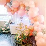 Elegant bridal shower setup with peach balloon arch, white bathtub, gold bar cart, and fresh floral arrangements