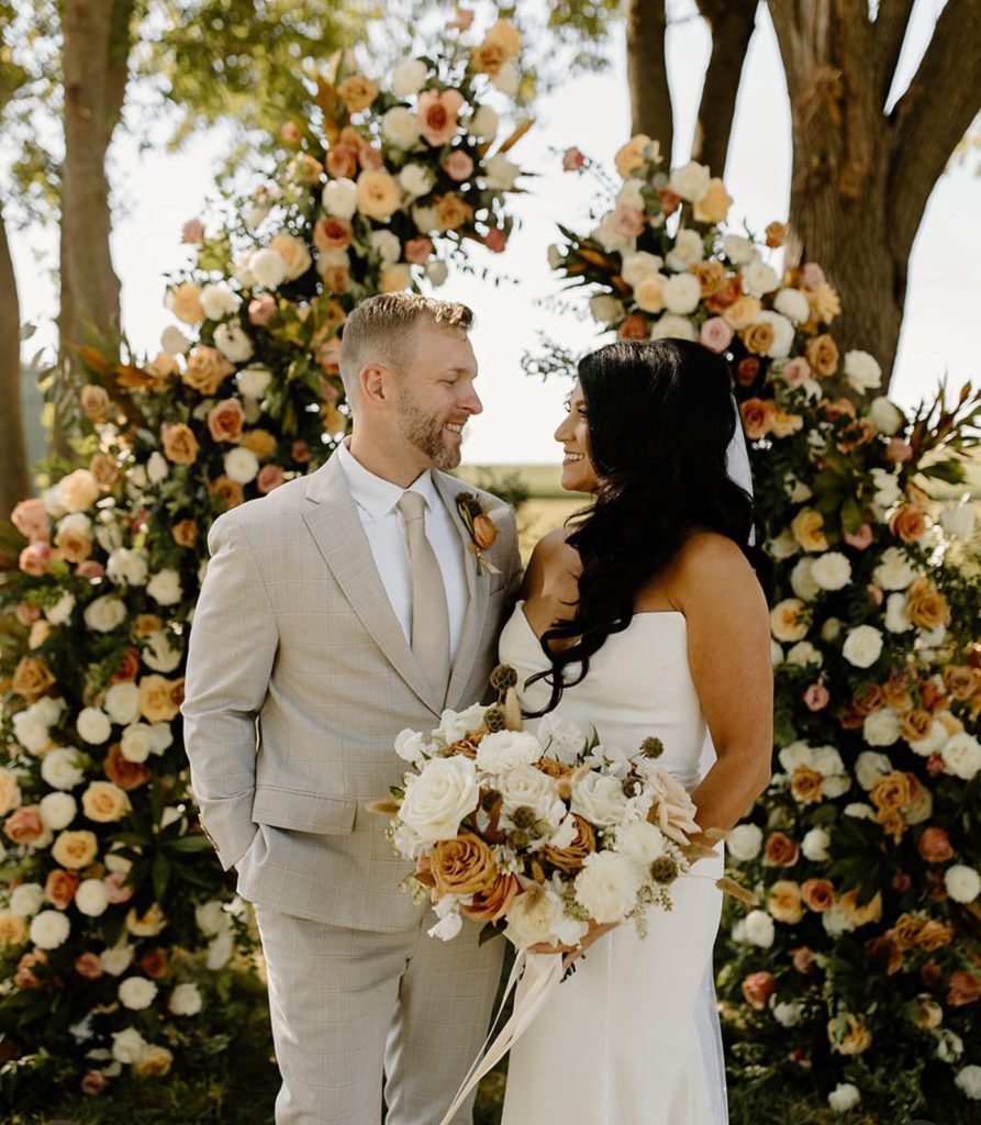 Bride and groom stand together before a vibrant floral ceremony arch featuring peachy and cream blooms in an outdoor Iowa setting