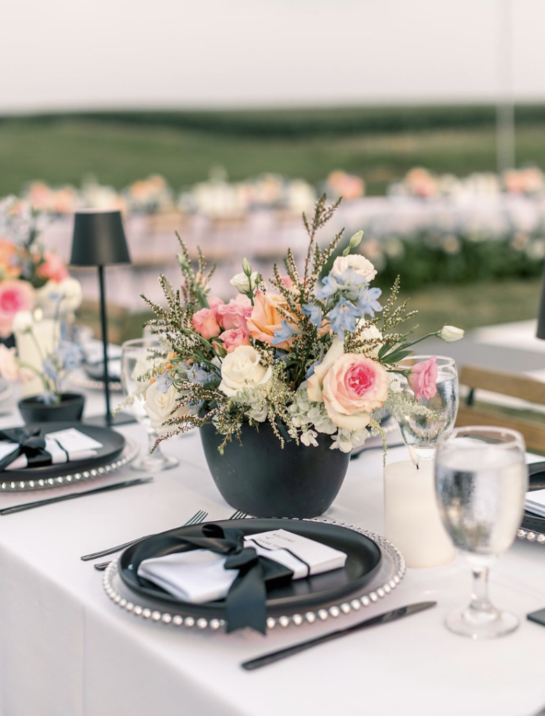 Outdoor reception table with pastel floral centerpiece in black vase, black plates with ribbon-tied napkins, and pearl-trimmed chargers