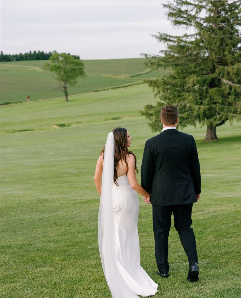Bride and groom holding hands walking across golf course fairway with rolling hills in background