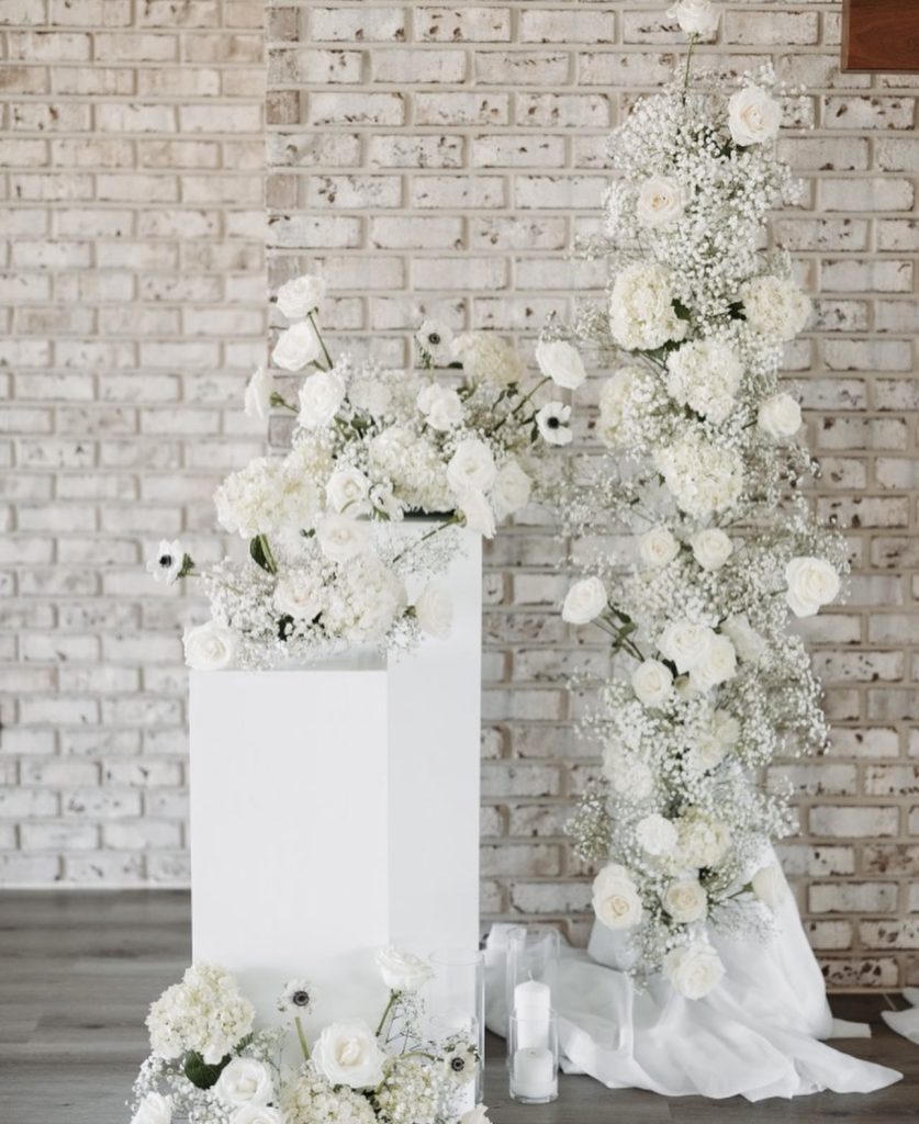 White floral ceremony arch with blooms and baby's breath against whitewashed brick wall