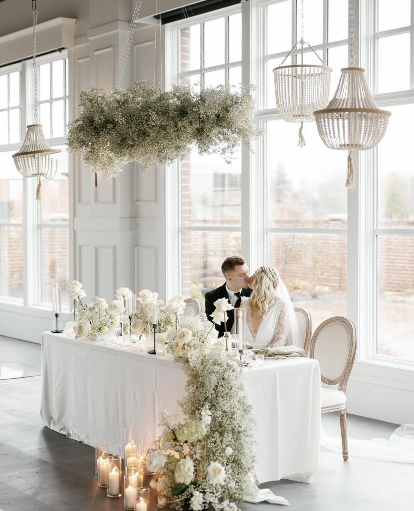 Newlyweds share a kiss at sweetheart table decorated with white blooms, greenery, and beaded chandeliers