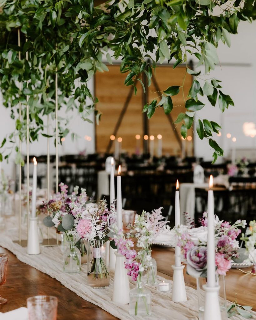 Reception table adorned with pink and white floral arrangements in glass vases, white taper candles, and cascading greenery overhead