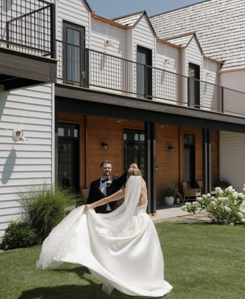 Groom twirls bride in flowing white gown outside modern venue with balcony