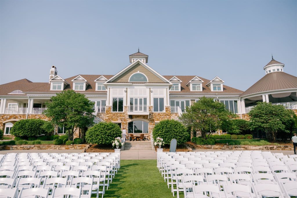 Outdoor wedding ceremony setup with white chairs arranged on lawn facing elegant stone and white building with cupola