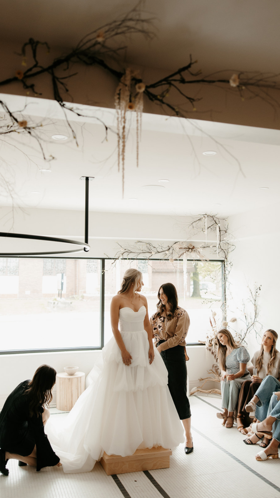 Bride in ballgown tries on dress at bridal boutique fitting with attendees watching under branch installations