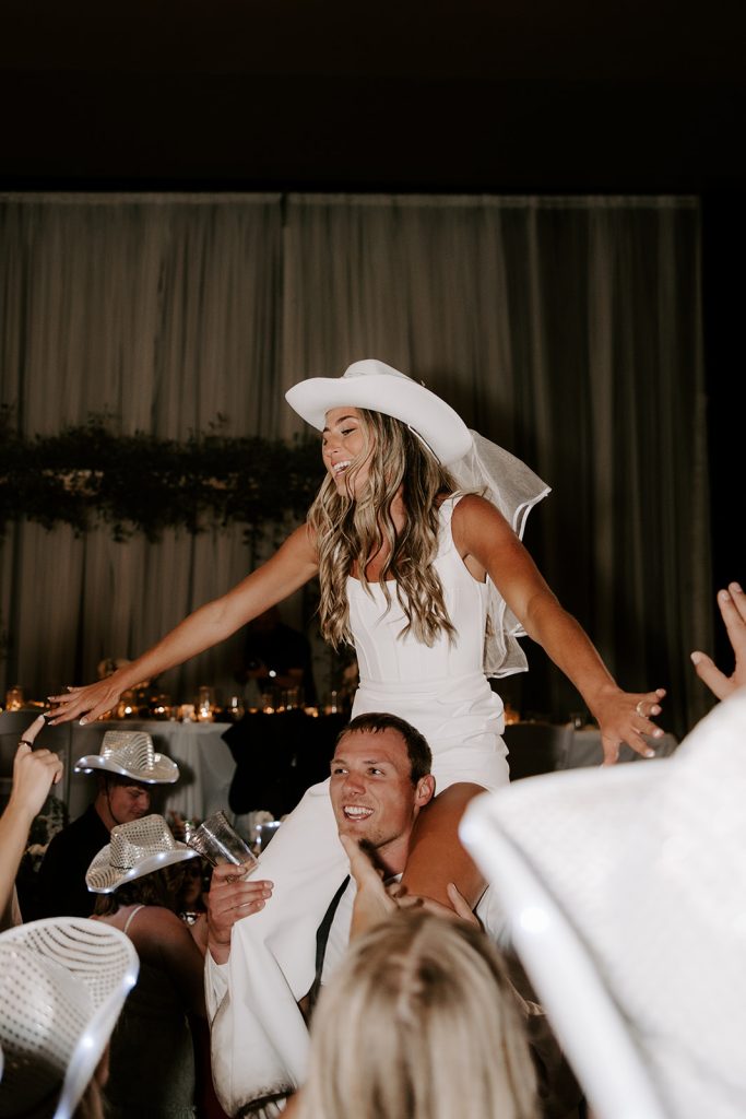 Bride wearing white cowboy hat celebrates on groom's shoulders during reception with guests in matching hats