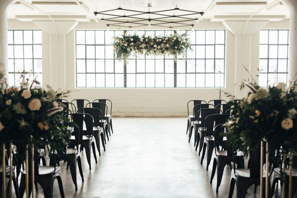 Industrial ceremony space with black chairs flanking aisle, overhead floral installation, and large windows