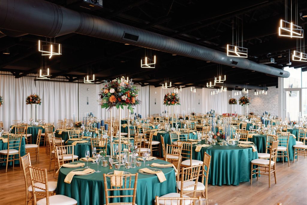 Modern industrial reception venue with teal tablecloths, gold chiavari chairs, tall floral centerpieces, and geometric pendant lighting
