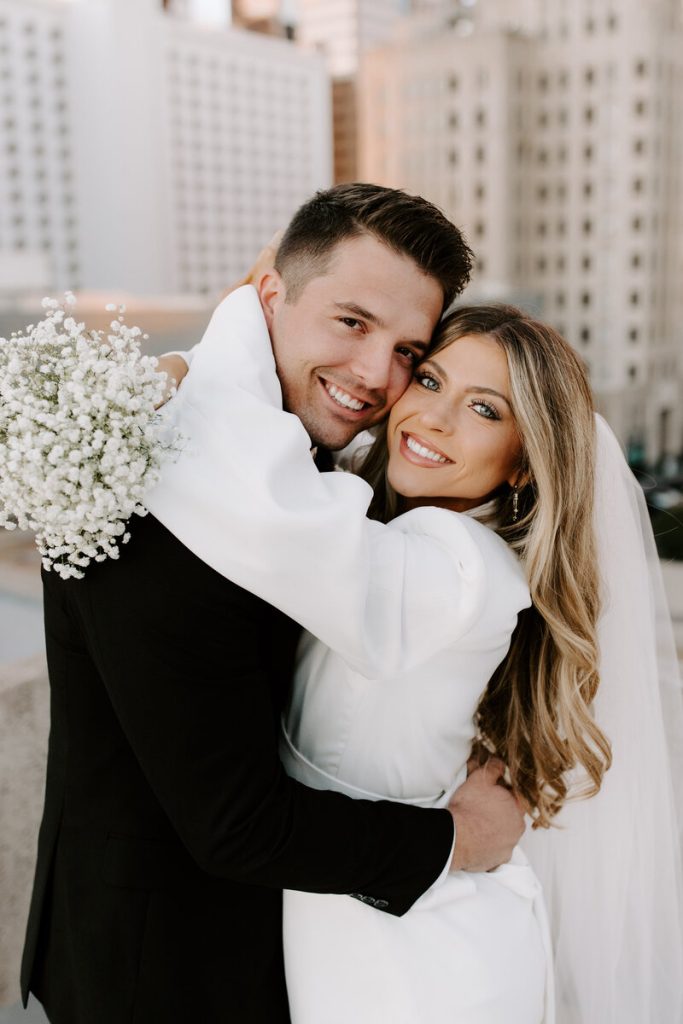 Newlyweds smiling and embracing with bouquet of white blooms against urban backdrop