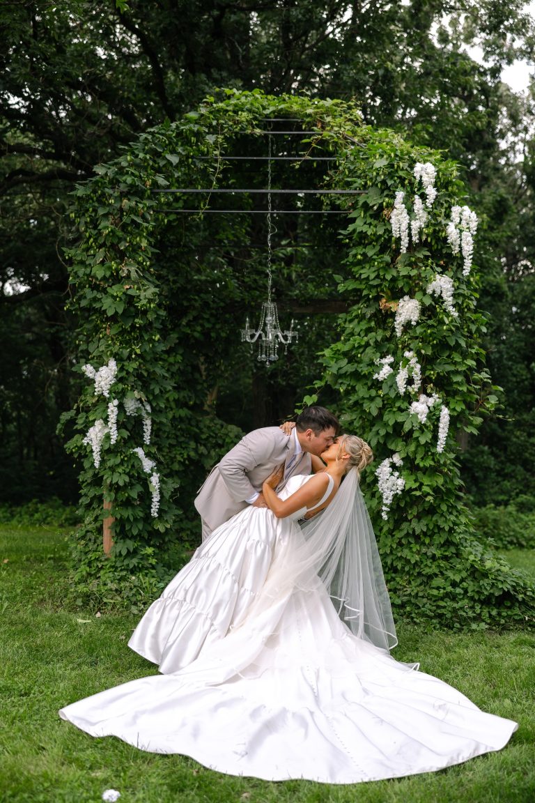 Couple kissing under vine-covered arbor decorated with cascading white blooms and hanging chandelier