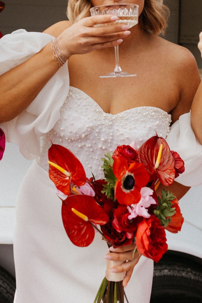 Bride in pearl-embellished gown holds vibrant red tropical bouquet and cocktail glass
