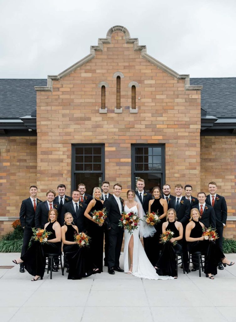 Wedding party in black attire with orange bouquets posed in front of historic brick building with arched windows