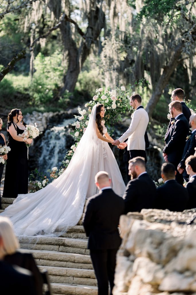 Outdoor ceremony with bride and groom exchanging vows beneath floral arch near waterfall