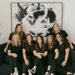 Team of seven women in matching black uniforms posing together on a couch in a modern office