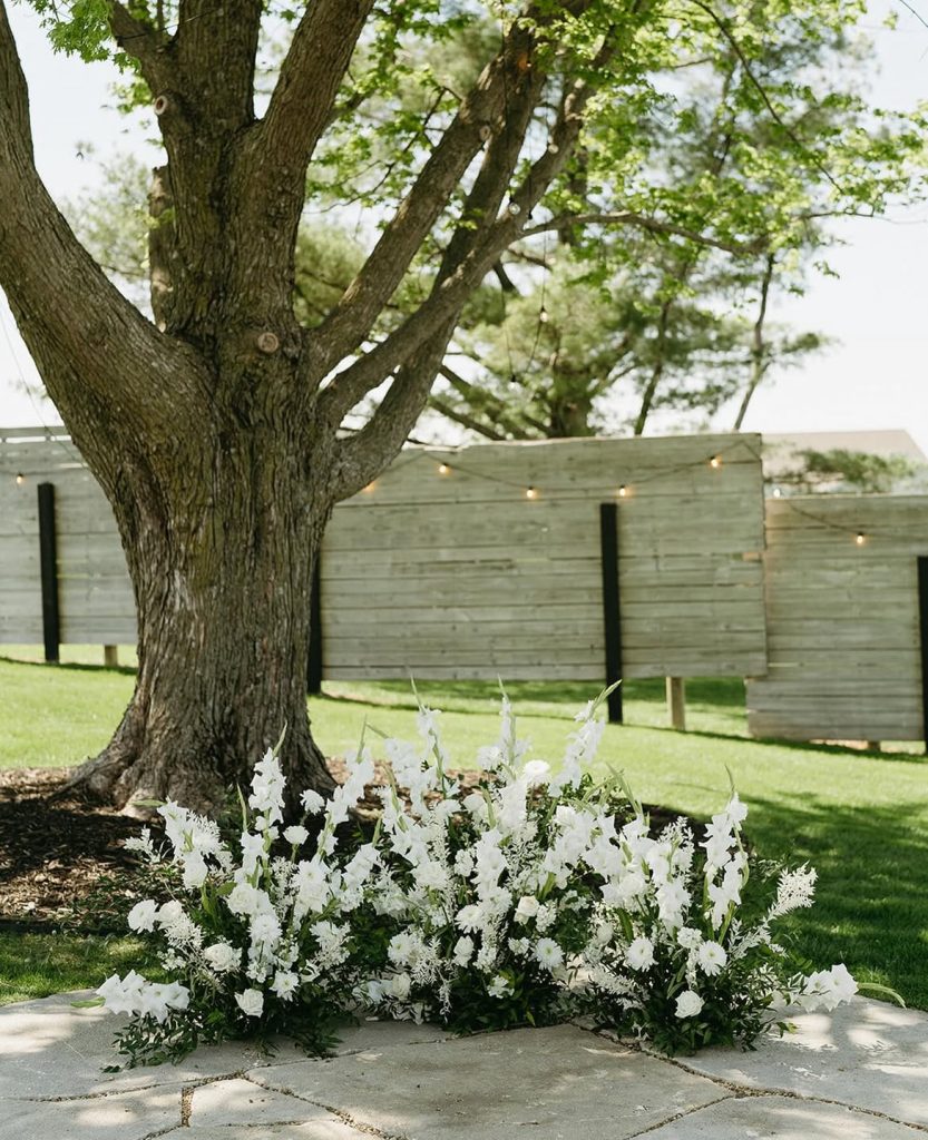 Lush white floral arrangement with greenery at base of large tree on outdoor ceremony patio