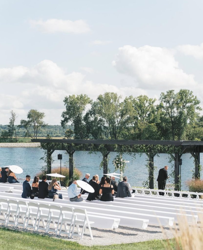 Lakeside ceremony with white chairs facing flower-adorned pergola, guests seated under cloudy sky