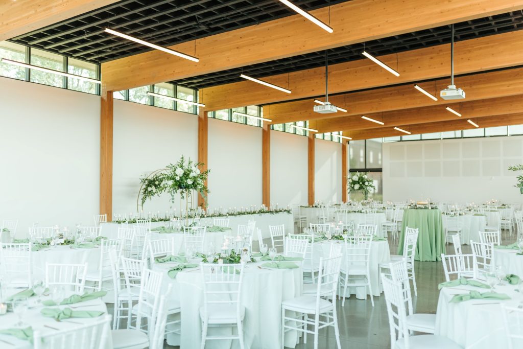 Modern reception hall with white chiavari chairs, sage green linens, and greenery centerpieces under warm wood beam ceiling