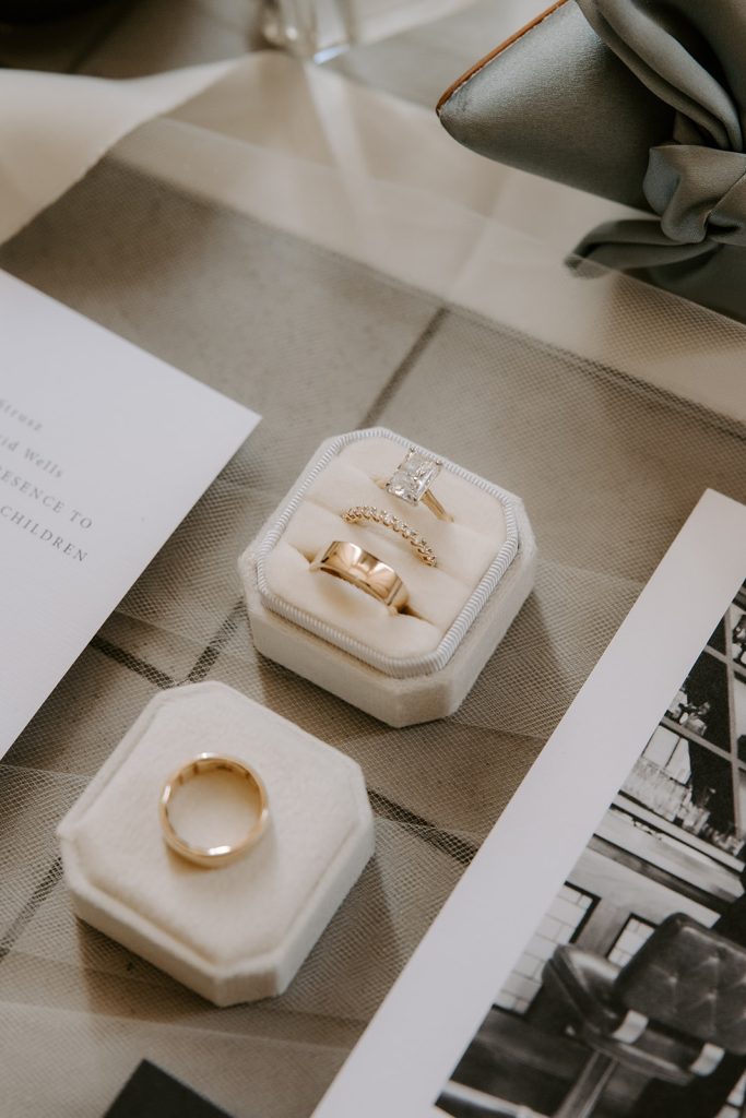 Gold wedding bands and diamond engagement ring displayed in velvet ring boxes with invitation suite