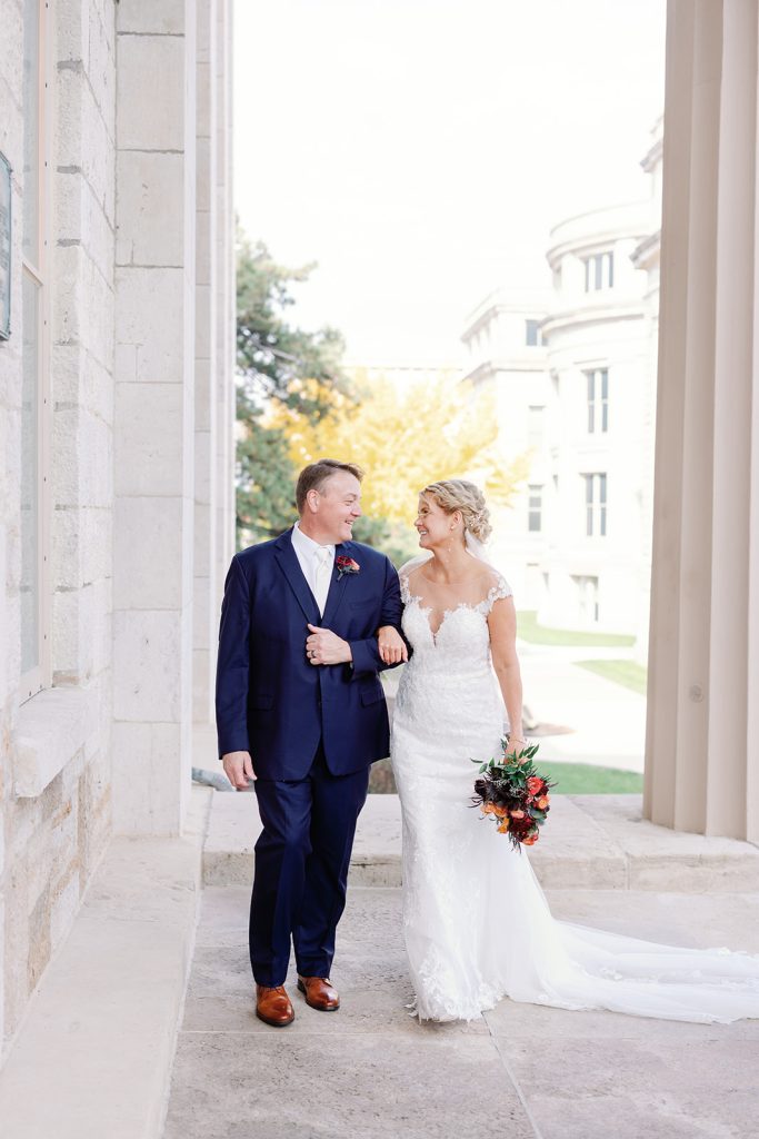 Bride in lace gown and groom in navy suit walk arm-in-arm through columned archway holding autumn bouquet