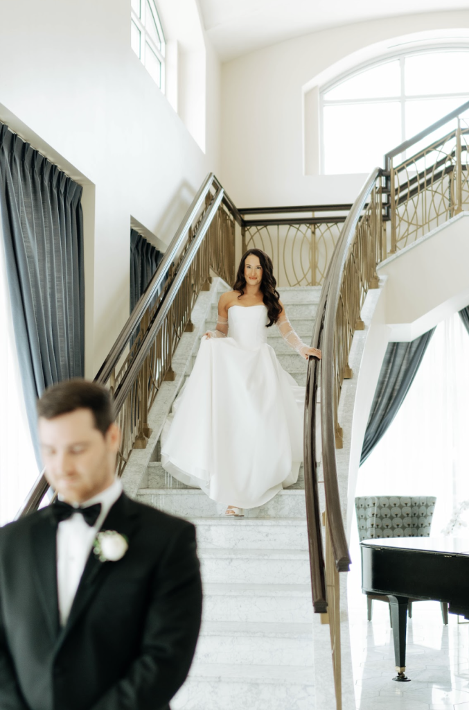 Bride in flowing white gown descends grand staircase during first look as groom waits below