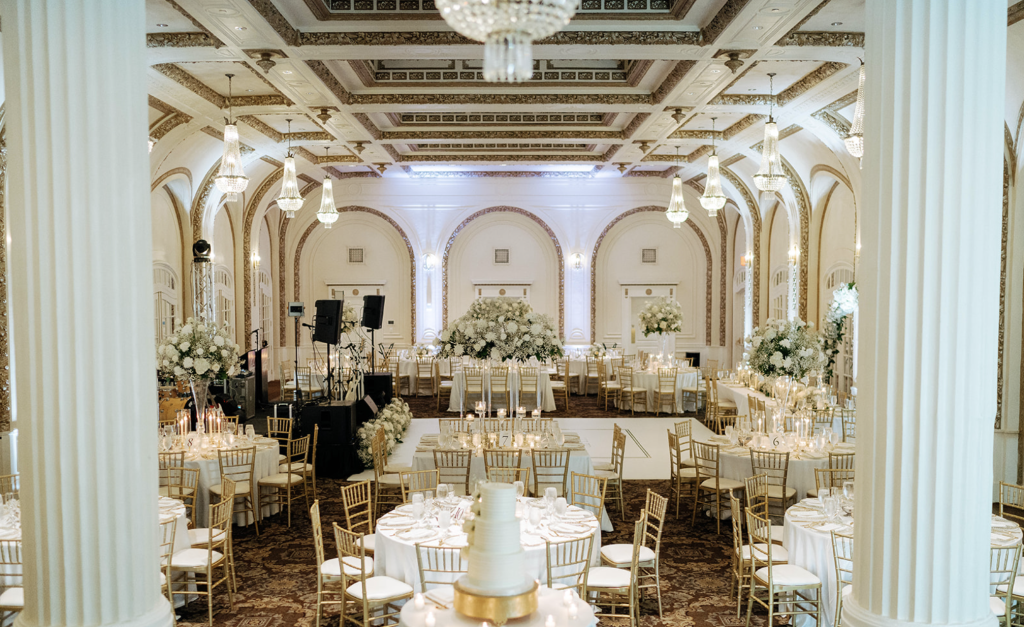 Elegant ballroom with gold chiavari chairs, white linens, tall floral centerpieces, and ornate coffered ceiling with crystal chandelier