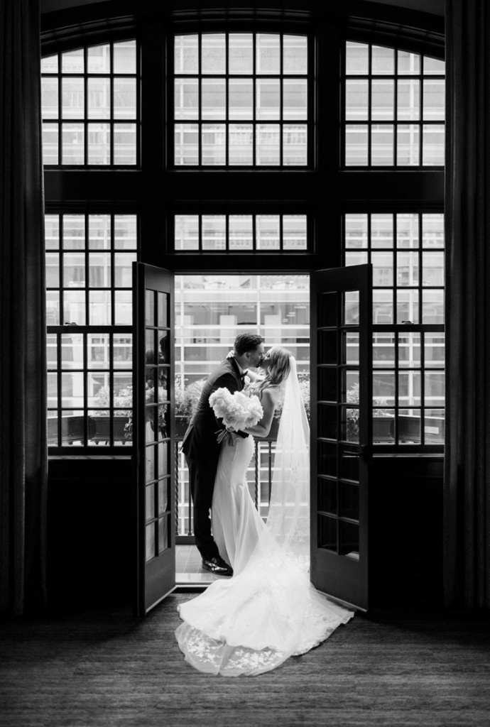 Bride and groom kiss in doorway framed by black French doors in dramatic black and white portrait
