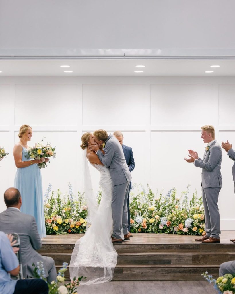 Newlyweds share first kiss during indoor ceremony with colorful blooms and wedding party