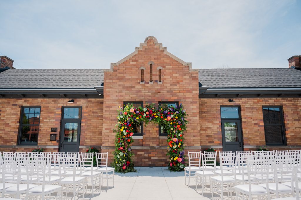 Outdoor ceremony setup with colorful floral arch and white chairs facing brick building