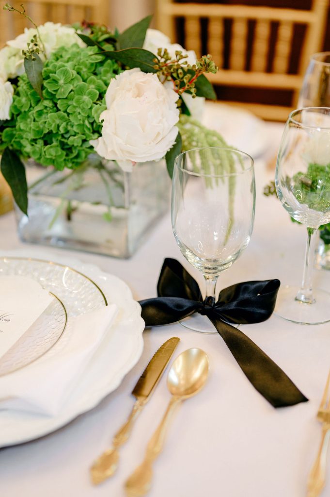 Elegant place setting with white and green floral centerpiece, gold flatware, and black ribbon detail on stemware