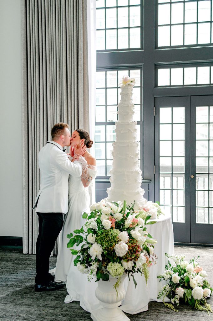 Couple kisses beside towering white textured wedding cake adorned with lush floral arrangements