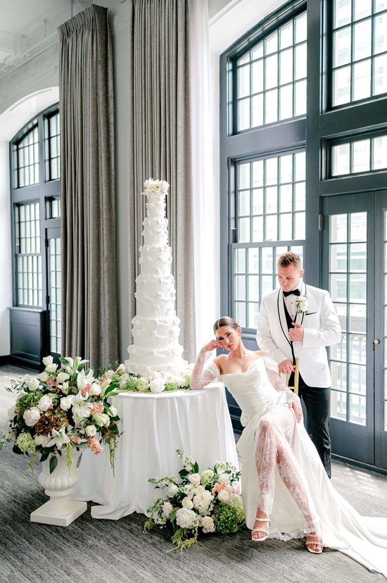Newlyweds pose beside elegant white textured cake and floral arrangements near tall windows