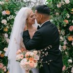 Newlyweds embrace before a lush floral arch adorned with peach and blush blooms