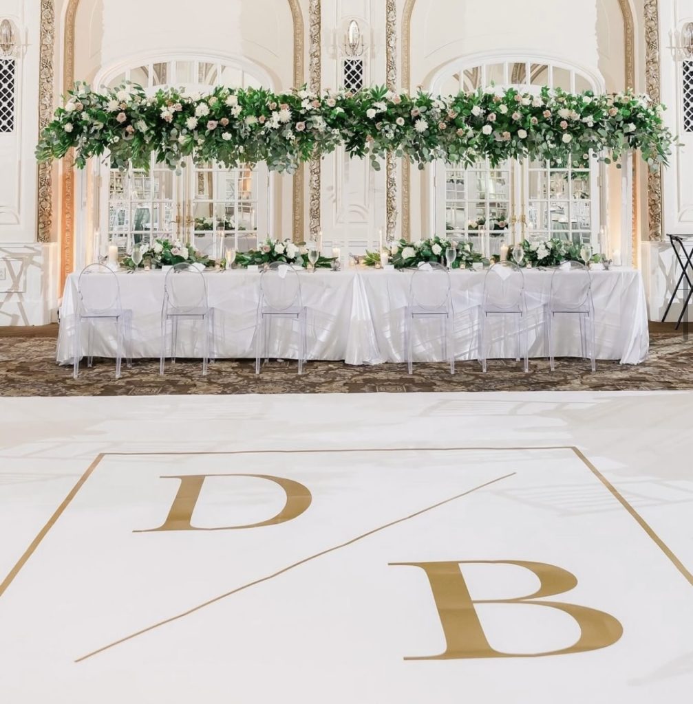 Elegant head table with lush overhead greenery installation and clear ghost chairs in ornate ballroom