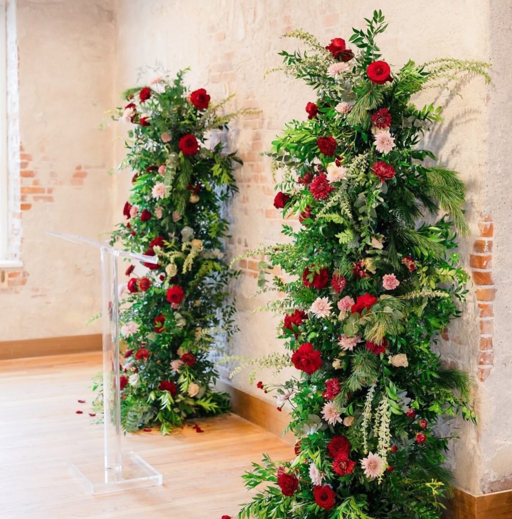 Lush floral arch with red and pink blooms adorning ceremony entrance against exposed brick wall
