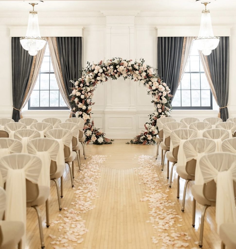 Indoor ceremony space with circular floral arch and rose petal-strewn aisle flanked by cream chairs