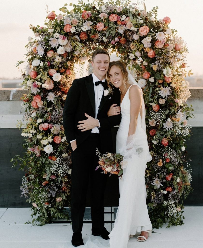 Couple posing beneath lush floral arch with coral and white blooms
