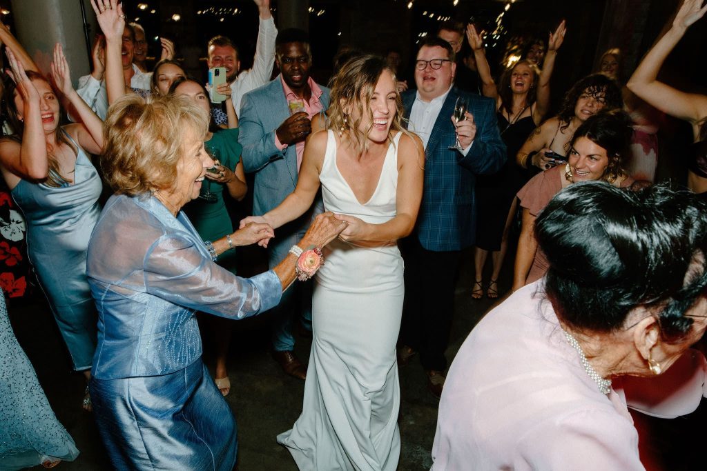 Bride in elegant white gown dances joyfully with guests on the reception dance floor surrounded by celebrating family and friends