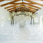 White wedding ceremony space with exposed wood beams, iron chandeliers, draped backdrop with flowering trees, and chiavari chairs