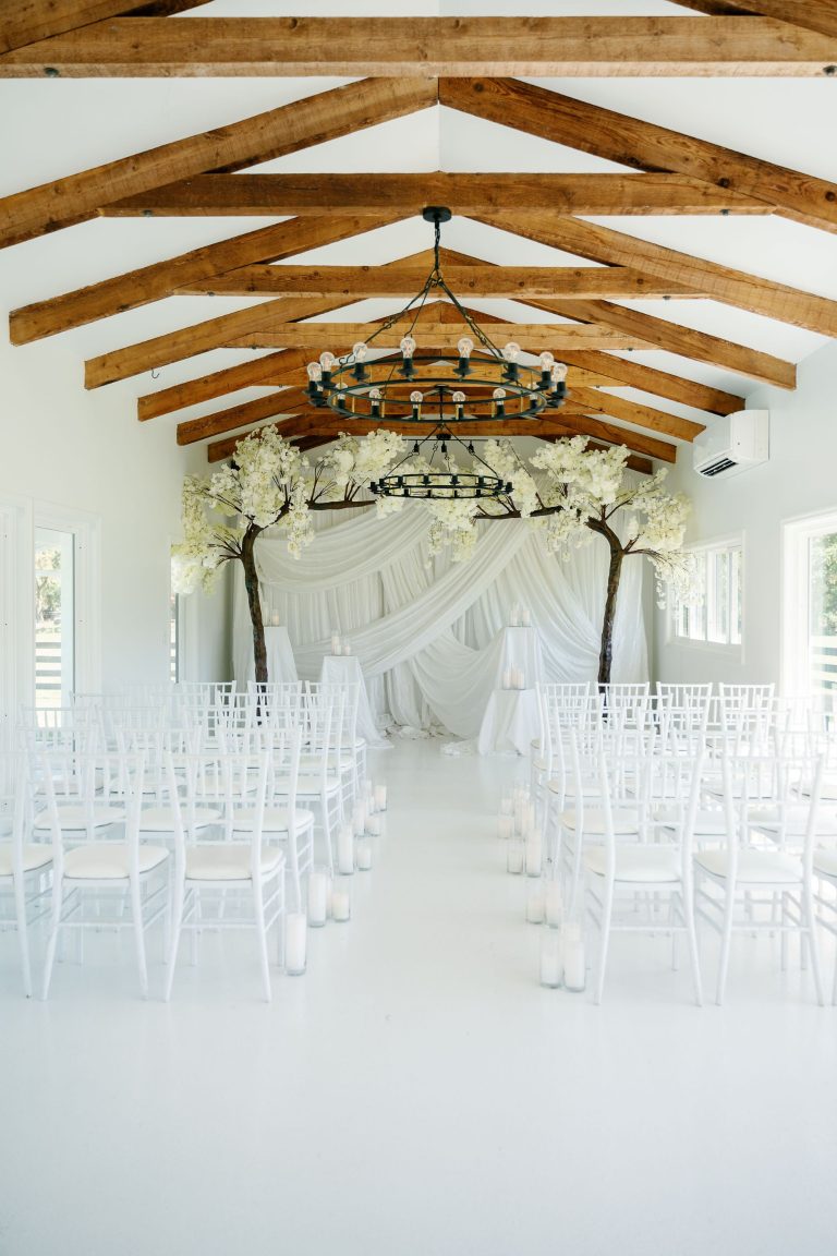 White wedding ceremony space with exposed wood beams, iron chandeliers, draped backdrop with flowering trees, and chiavari chairs