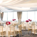 Elegant ballroom reception setup with gold chiavari chairs, draped ceiling, and pink floral centerpieces