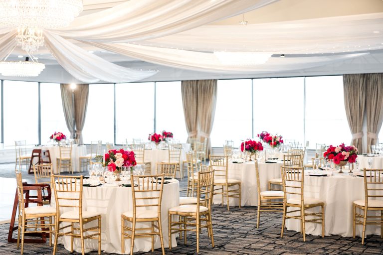 Elegant ballroom reception setup with gold chiavari chairs, draped ceiling, and pink floral centerpieces