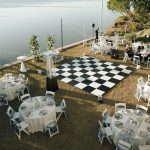 Waterfront wedding reception setup with checkered dance floor, white tables, and wooden dock ceremony area overlooking lake