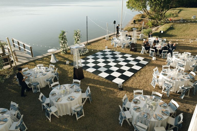Waterfront wedding reception setup with checkered dance floor, white tables, and wooden dock ceremony area overlooking lake