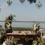 Wooden ceremony arbor adorned with white floral arrangements and crystal chandelier overlooking waterfront