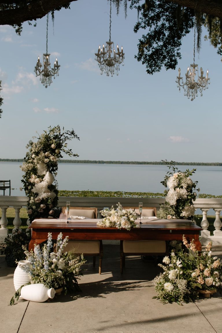 Wooden ceremony arbor adorned with white floral arrangements and crystal chandelier overlooking waterfront