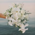 Cascading white bridal bouquet with greenery held against ocean backdrop at sunset