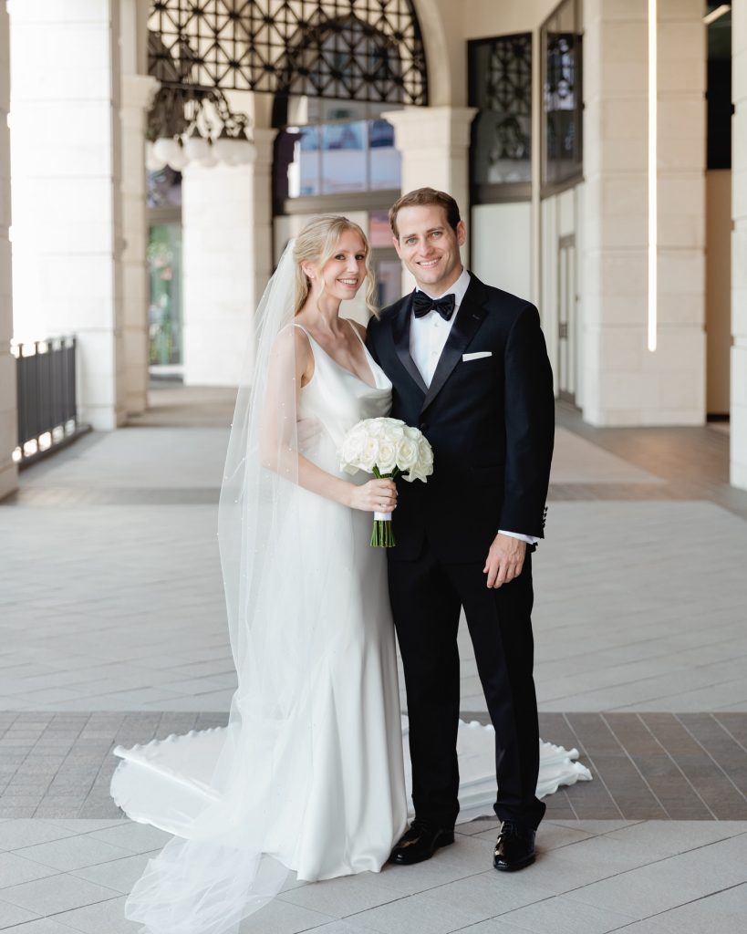 Bride in sleek satin gown and groom in black tuxedo pose with white bouquet under covered walkway