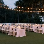 Outdoor wedding reception setup with gold chiavari chairs, white linens, and string lights hung over Spanish moss-draped oak trees at dusk