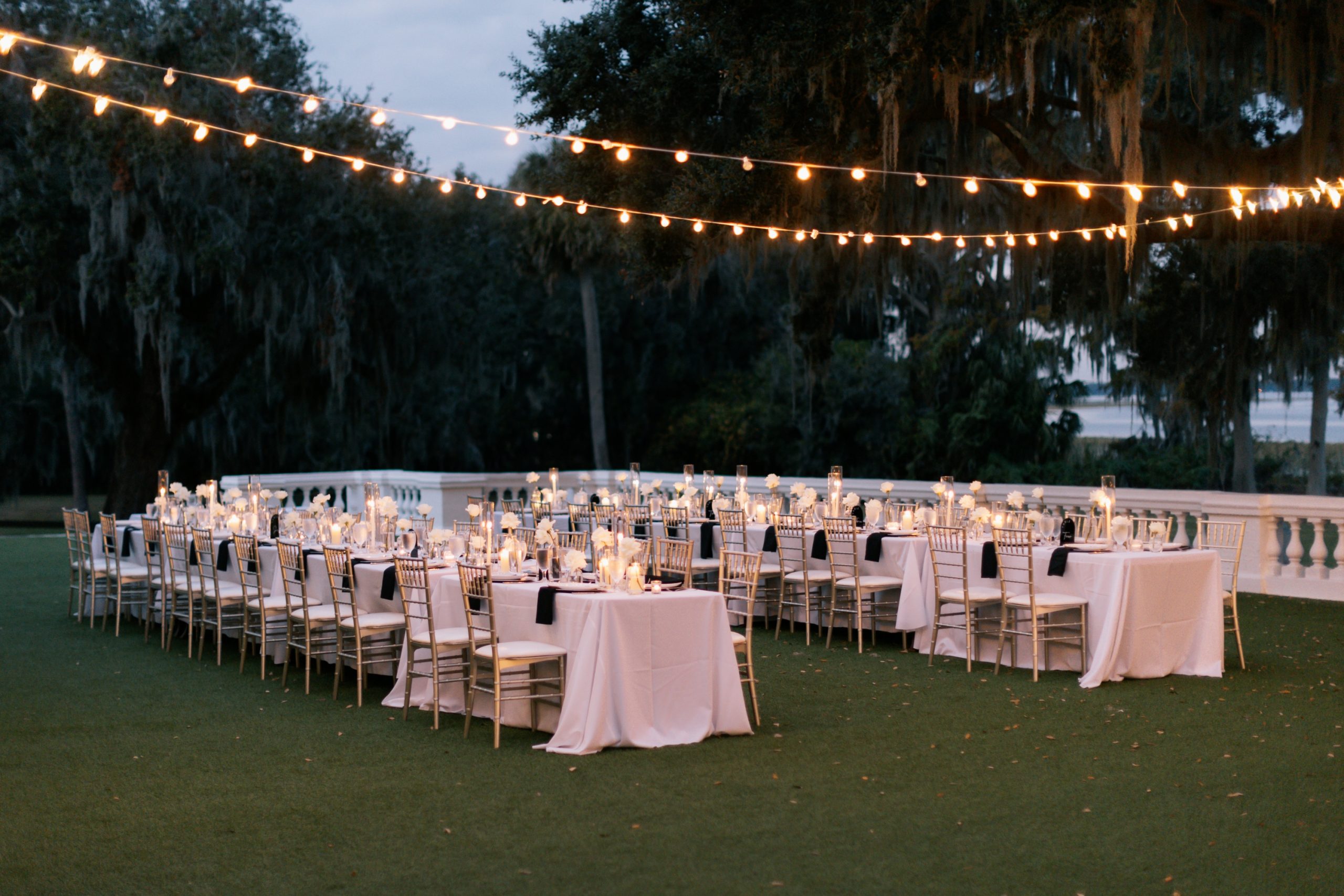 Outdoor wedding reception setup with gold chiavari chairs, white linens, and string lights hung over Spanish moss-draped oak trees at dusk